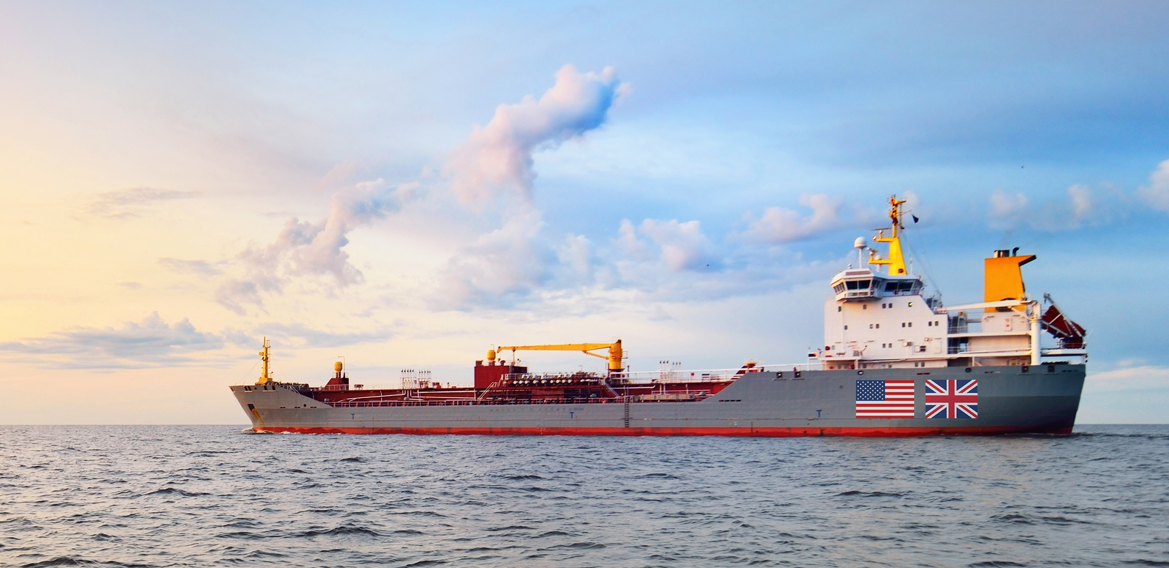 Cargo ship with UK and US flags sailing at dusk.